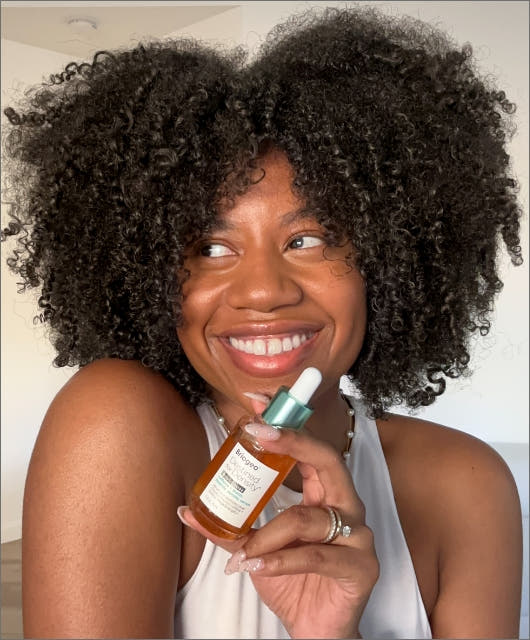 A woman with thick, black, very curly hair holding a bottle of Briogeo Density Serum. 